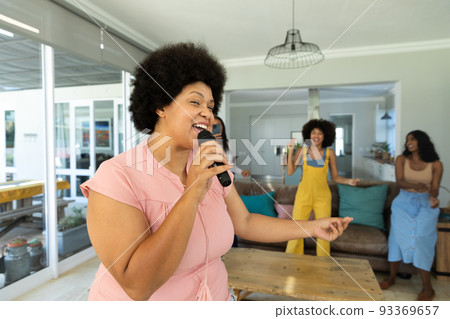 Happy biracial young woman with afro hair singing over microphone and friends dancing in background Happy biracial young woman with afro hair singing over microphone and friends dancing in background 93369657