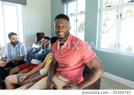 Portrait of smiling young man sitting with multiracial male friends on sofa against windows at home Portrait of smiling young man sitting with multiracial male friends on sofa against windows at home 93369745