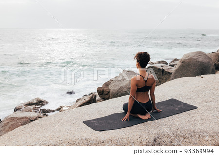 Back view of female in sportswear sitting in thunderbolt pose looking at ocean Back view of female in sportswear sitting in thunderbolt pose looking at ocean 93369994