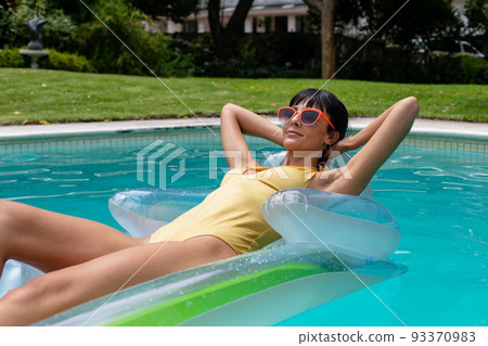 White young woman wearing swimsuit and sunglasses resting on inflatable raft in swimming pool 93370983