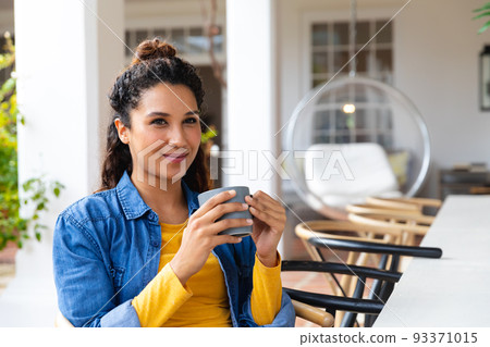 Happy biracial woman sitting on garden terrace drinking coffee outside house 93371015