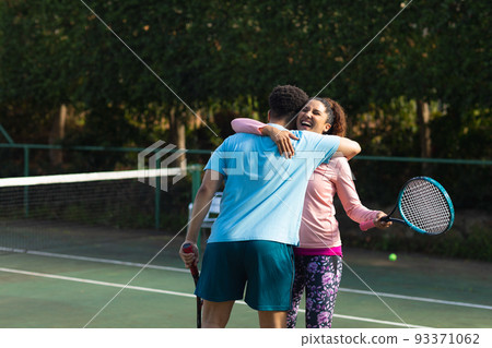 Smiling biracial couple playing tennis embracing on outdoor tennis court 93371062