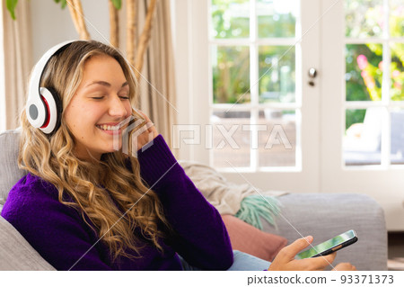 Happy caucasian woman on couch wearing headphones, listening to music smartphone in living room Happy caucasian woman on couch wearing headphones, listening to music smartphone in living room 93371373