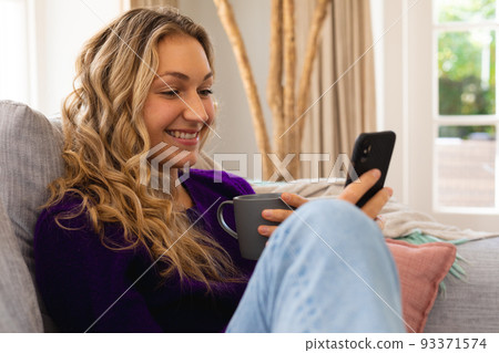 Smiling caucasian woman sitting on couch using smartphone in sunny living room 93371574