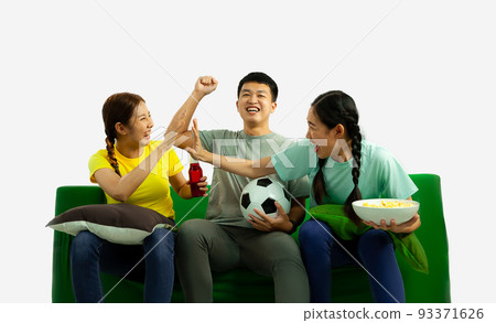 A group of young asian man and women holding football ball popcorn bowl sitting on green sofa cheering the soccer game at home on the white screen background. 93371626