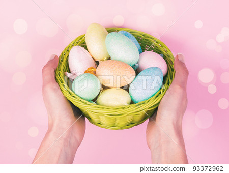 Woman hands holding wooden basket with colored ester eggs on pink background. Easter Concept 93372962