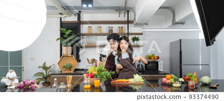 Asian couple spend time together in the kitchen. Young woman in apron cooking salad dish while his boyfriend recording vlog video for social blogger. Photography studio lights set up in the kitchen. 93374490