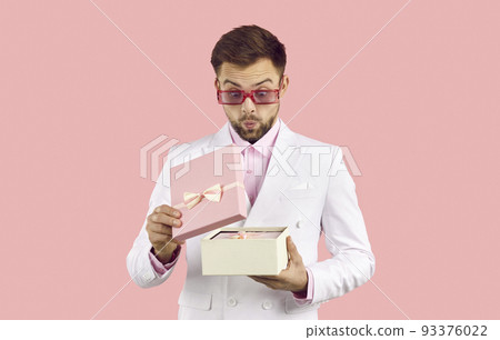 Funny surprised man with excited face looks at contents of gift box on pastel pink background. Funny surprised man with excited face looks at contents of gift box on pastel pink background. 93376022