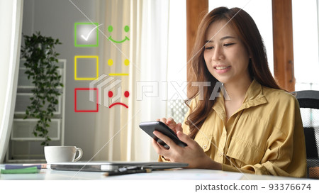 Beautiful young woman using mobile phone, giving satisfied rating with smiley face icon to evaluate product and service Beautiful young woman using mobile phone, giving satisfied rating with smiley face icon to evaluate product and service 93376674