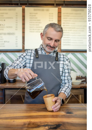 Barista pouring coffee to the coffee mug and looking contented Barista pouring coffee to the coffee mug and looking contented 93378028