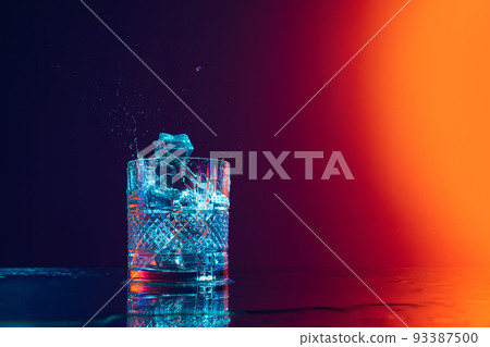 Ice cube falls into a transparent glass of water standing on mirror surface over gradient black orange background in neon light. Art, beauty, drinks Ice cube falls into a transparent glass of water standing on mirror surface over gradient black orange background in neon light. Art, beauty, drinks 93387500