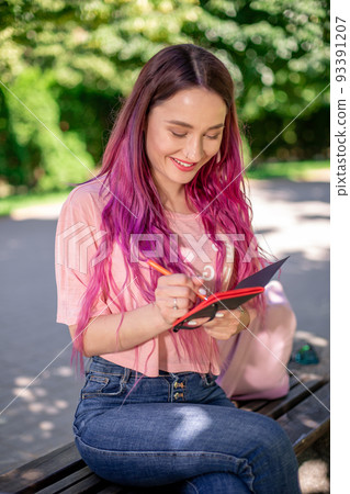 Woman writing in a notebook sitting on a wooden bench in the park. Girl working outdoors on portable computer, copy space. 93391207