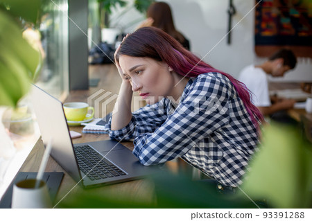 Side view of young businesswoman sitting at table in coffee shop. On table cup of coffee and laptop. In background white wall and window. 93391288