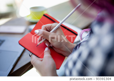 Positive female student with pink hair writing down funny story in notebook sitting in coffee shop with cup of cappuccino and laptop. Positive female student with pink hair writing down funny story in notebook sitting in coffee shop with cup of cappuccino and laptop. 93391304