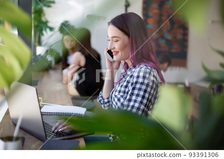 Side view of young businesswoman sitting at table in coffee shop. On table cup of coffee and laptop. In background white wall and window. Side view of young businesswoman sitting at table in coffee shop. On table cup of coffee and laptop. In background white wall and window. 93391306