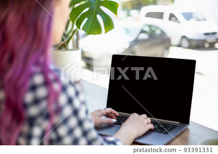 Back view of a young pink hair woman keyboarding on laptop computer with blank copy space screen while sitting in cafe. 93391331