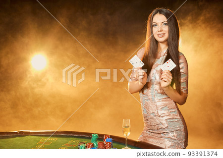 Gorgeous young woman in evening dress standing near poker table with glass of champagne Gorgeous young woman in evening dress standing near poker table with glass of champagne 93391374