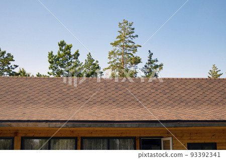 The roof of the wooden house is covered with shingles. Against the background of the blue sky and coniferous trees The roof of the wooden house is covered with shingles. Against the background of the blue sky and coniferous trees 93392341
