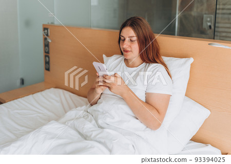 Beautiful young woman reading text message on her mobile phone in the Bed. Check social networks, send sms. The girl is wearing a white T-shirt. Beautiful young woman reading text message on her mobile phone in the Bed. Check social networks, send sms. The girl is wearing a white T-shirt. 93394025
