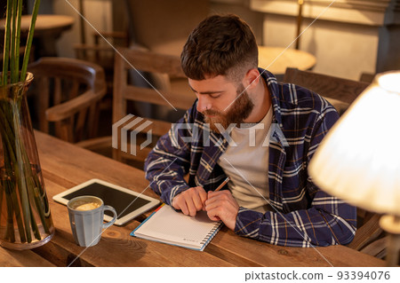 Young bearded businessman sits in cafe, home at table and writes in notebook, near lies tablet computer with black screen. Man is working, studying. 93394076