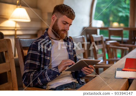 Freelancer dressed in casual outfit focus on reading news and looking on digital tablet while sitting in cozy urban cafe. 93394147