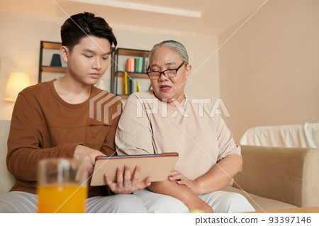 Asian young man installing applications on tablet computer of grandmother so she could call her friends Asian young man installing applications on tablet computer of grandmother so she could call her friends 93397146