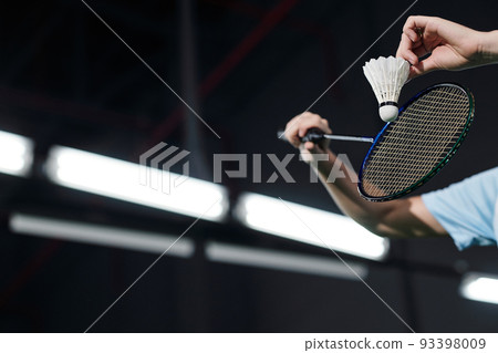 Hands of male player holding racket and hitting shuttlecock on dark badminton indoor court 93398009