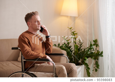 Serious young man with disability sitting in wheelchair and talking on phone 93398083