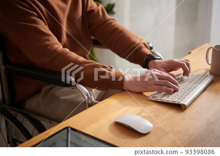 Closeup image of young man sitting in wheelchair working on laptop at his desk Closeup image of young man sitting in wheelchair working on laptop at his desk 93398086