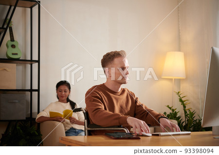 Serious young man in wheelchair working on computer, his girlfriend reading book in background 93398094