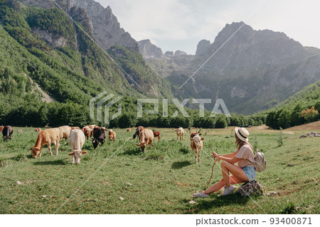 Young woman sitting and relaxing on alpine mountain looking the cow eating a grass in the summer. Travel, Lifestyle Concept. Beautiful woman enjoys views of the alpine village in the Alps mountains Young woman sitting and relaxing on alpine mountain looking the cow eating a grass in the summer. Travel, Lifestyle Concept. Beautiful woman enjoys views of the alpine village in the Alps mountains 93400871