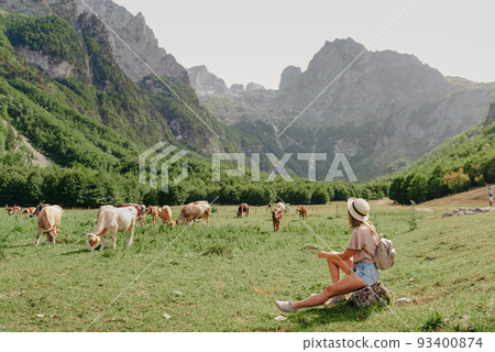 Young woman sitting and relaxing on alpine mountain looking the cow eating a grass in the summer. Travel, Lifestyle Concept. Beautiful woman enjoys views of the alpine village in the Alps mountains Young woman sitting and relaxing on alpine mountain looking the cow eating a grass in the summer. Travel, Lifestyle Concept. Beautiful woman enjoys views of the alpine village in the Alps mountains 93400874