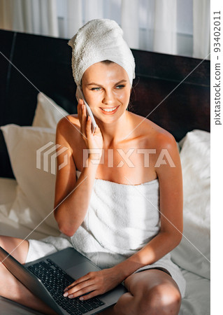 Pretty woman wrapped in bath towel lying on bed with laptop on knees and holding smartphone in hands. Lifestyles with modern technology. Young girl with a laptop talking on the phone. Pretty woman wrapped in bath towel lying on bed with laptop on knees and holding smartphone in hands. Lifestyles with modern technology. Young girl with a laptop talking on the phone. 93402011