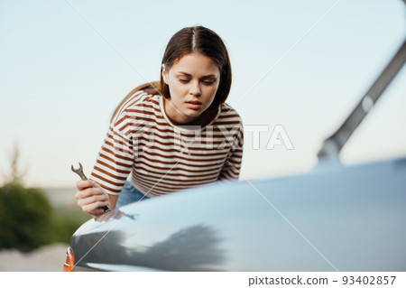 A woman with a wrench looks sadly and sadly under the open hood of her car and can't fix it from a roadside breakdown while traveling alone A woman with a wrench looks sadly and sadly under the open hood of her car and can't fix it from a roadside breakdown while traveling alone 93402857