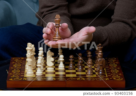 The hands of a white-skinned man carefully hold the black chess piece of the king above the board, showing it off. 93403156