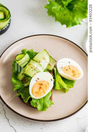 Ready to eat sandwich with rye bread, lettuce, cucumber, boiled egg and flax seeds on a plate on the table. Healthy eating on a diet. Vertical view. Close-up Ready to eat sandwich with rye bread, lettuce, cucumber, boiled egg and flax seeds on a plate on the table. Healthy eating on a diet. Vertical view. Close-up 93403226