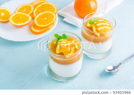Orange panna cotta with mint leaf in glasses and orange slices on a plate on a light table. Sweet Italian dessert Orange panna cotta with mint leaf in glasses and orange slices on a plate on a light table. Sweet Italian dessert 93403966