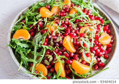 Fruit vitamin salad of pomegranate, tangerine, arugula and pumpkin seeds in a plate on the table. Organic vegetarian food. Close-up 93404014