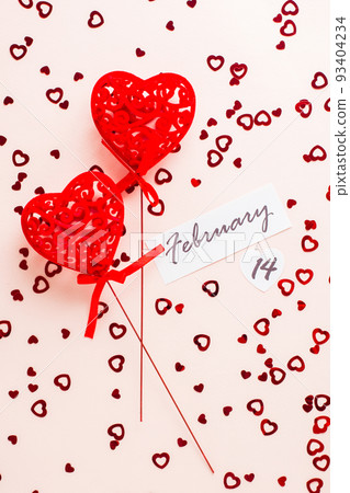 Valentine's Day. Date of February 14 and two curly red hearts on a pink background strewn with hearts. Vertical and top view Valentine's Day. Date of February 14 and two curly red hearts on a pink background strewn with hearts. Vertical and top view 93404234