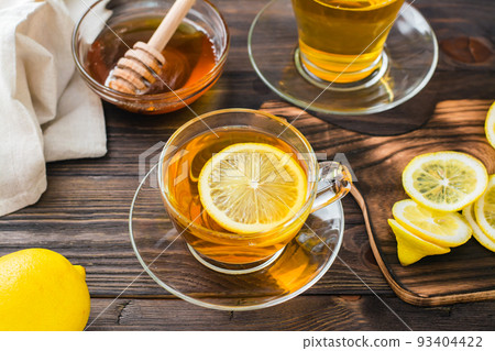 A cup of tea with a slice of lemon and a bowl of honey on a wooden table. Vitamin warming drink. Close-up A cup of tea with a slice of lemon and a bowl of honey on a wooden table. Vitamin warming drink. Close-up 93404422