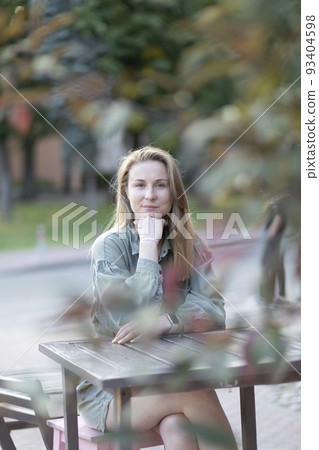 Sustainable, young smiling lovely European slav female blondie is sitting at cafe table outside , wears green - khaki denim Relaxed in summer day time. Vertical plane. Sustainable, young smiling lovely European slav female blondie is sitting at cafe table outside , wears green - khaki denim Relaxed in summer day time. Vertical plane. 93404598