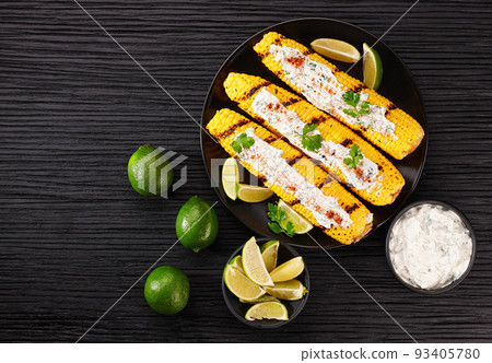 Grilled corn grilled on fire with cream sauce (mexican elotes), hot chili pepper, lime. On a dark table, copy space for text. 93405780