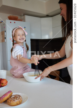 Mother and daughter preparing a sweet cake using flour, milk, sitting on chairs at a table in a modern kitchen. Girl holding a whisk, stirring eggs in a bowl, preparing pancake dough with her mom. 93405820