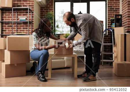 Diverse couple preparing boxes with sticky tape to move in first house bought on mortgage loan, relocating property together. Packing up furniture with adhesive roller in new apartment. Diverse couple preparing boxes with sticky tape to move in first house bought on mortgage loan, relocating property together. Packing up furniture with adhesive roller in new apartment. 93411032