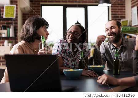Positive diverse friends at home laughing and talking together while watching movies on laptop. Happy smiling people in living room enjoying fun leisure activity while having snacks. 93411080