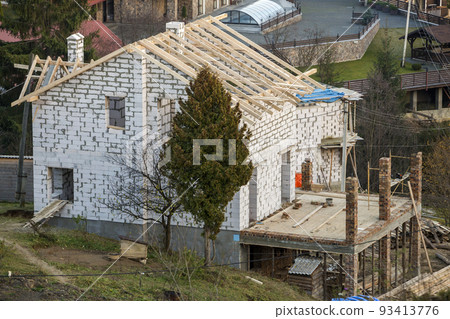 Two-storied cottage under construction. Walls of hollow foam insulation blocksand with window openings,roof beams frame and high chimneys, roofing underlayment. 93413776