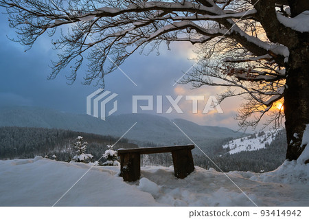 Moody winter landscape with dark bare tree and small wooden bench on covered with fresh fallen snow field in wintry mountains on cold gloomy evening. Moody winter landscape with dark bare tree and small wooden bench on covered with fresh fallen snow field in wintry mountains on cold gloomy evening. 93414942