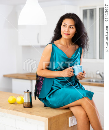 Woman in nightie sitting on table at home and drinking tea Woman in nightie sitting on table at home and drinking tea 93415697