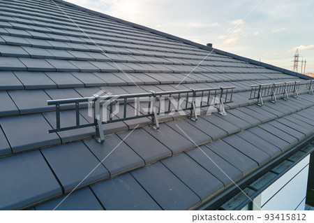 Closeup snow guard for safety in winter on house roof top covered with ceramic shingles. Tiled covering of building 93415812