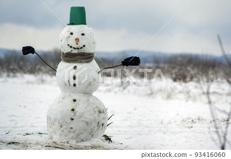 Big smiling snowman with bucket hat, scarf and gloves on white snowy field winter landscape, blurred black trees and blue sky copy space background. . Merry Christmas and happy new year greeting card. Big smiling snowman with bucket hat, scarf and gloves on white snowy field winter landscape, blurred black trees and blue sky copy space background. . Merry Christmas and happy new year greeting card. 93416060